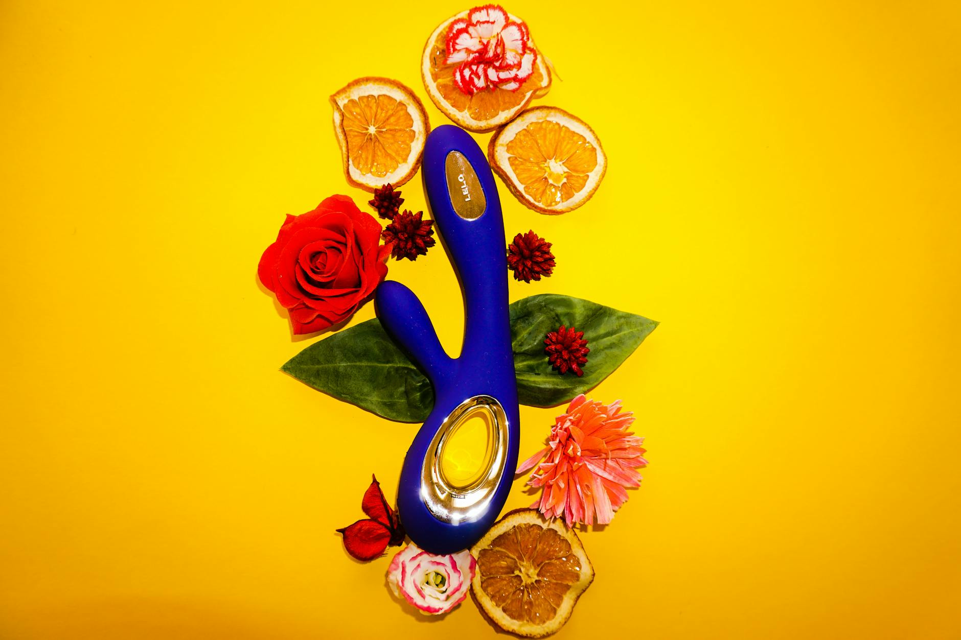 A blue vibrator surrounded by flowers and citrus on a bright yellow background, symbolizing renewed pleasure and self-connection